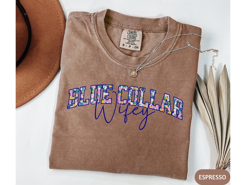 May include: A brown t-shirt with the text "BLUE COLLAR Wifey" in a floral design. A silver necklace with a pendant is on the shirt. A brown hat is visible in the upper left corner. The shirt is a Comfort Colors brand.
