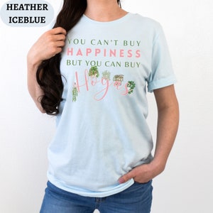 May include: A light blue t-shirt with the text "You can't buy happiness but you can buy Hoyas" in pink lettering. The text is surrounded by illustrations of potted plants.