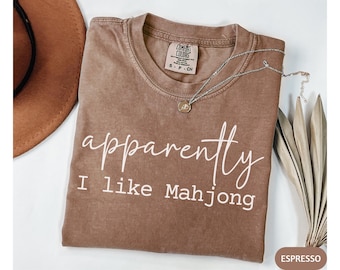 May include: Espresso-colored t-shirt with the text "apparently I like Mahjong" in white script. A silver necklace with a small pendant rests on the shirt. A brown hat is visible in the upper left corner.