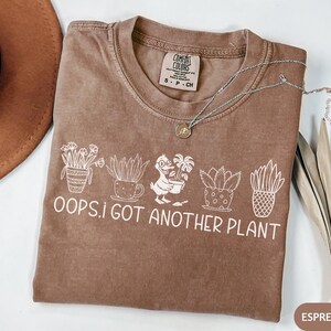 May include: Espresso-colored t-shirt with the text "OOPS, I GOT ANOTHER PLANT" in white. The shirt features a line of illustrated plants. A silver necklace with a small pendant is on the shirt. A brown hat is in the background.