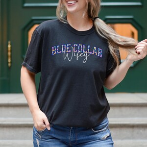 May include: Black t-shirt featuring the text "BLUE COLLAR Wifey" in a colourful, floral-patterned design. The shirt has a crew neck and short sleeves. The model is wearing blue jeans.