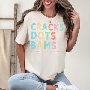 May include: Ivory-colored t-shirt with the words "CRACKS DOTS BAMS" in colorful, pastel letters. Below the text is the phrase "If you know, you know." The shirt is a casual style.