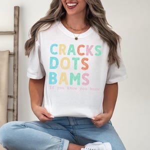 May include: White t-shirt with the words "CRACKS DOTS BAMS" in colorful, pastel letters. Below the words is the phrase "If you know you know". The shirt is a casual, everyday wear item.