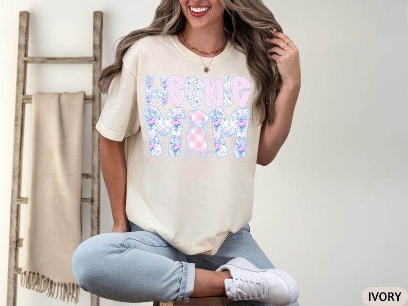 K&ouml;nnte beinhalten: Elfenbeinfarbenes T-Shirt mit dem Text "Home Mom" in einem floralen und Vichy-Muster. Das Design zeigt einen Dackel. Das Shirt hat eine lockere Passform. Dazu werden blaue Jeans und wei&szlig;e Turnschuhe getragen.