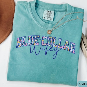 May include: Seafoam green t-shirt with the text "BLUE COLLAR Wifey" in a floral design. A gold necklace with a pendant rests on the shirt. The shirt is a Comfort Colors brand, size S-CH.