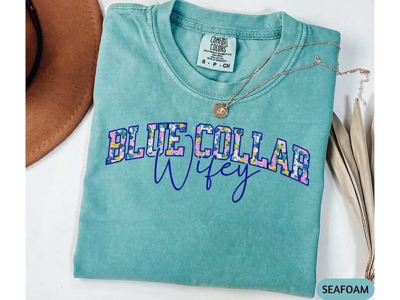 May include: Seafoam green t-shirt with the text "BLUE COLLAR Wifey" in a floral design. A gold necklace with a pendant rests on the shirt. The shirt is a Comfort Colors brand, size S-CH.