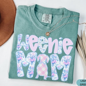 K&ouml;nnte beinhalten: Hellgr&uuml;nes T-Shirt mit den Worten "Weenie Mom" in einem Blumen- und Vichy-Muster. Eine Dackel-Grafik befindet sich in der Mitte. Eine goldene Halskette und ein brauner Hut sind im Hintergrund.