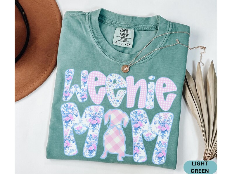 K&ouml;nnte beinhalten: Hellgr&uuml;nes T-Shirt mit den Worten "Weenie Mom" in einem Blumen- und Vichy-Muster. Eine Dackel-Grafik befindet sich in der Mitte. Eine goldene Halskette und ein brauner Hut sind im Hintergrund.