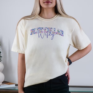 May include: Ivory t-shirt featuring the text "BLUE COLLAR WIFEY" in a floral design. The shirt has a relaxed fit and short sleeves. The text is in a blue and floral design.