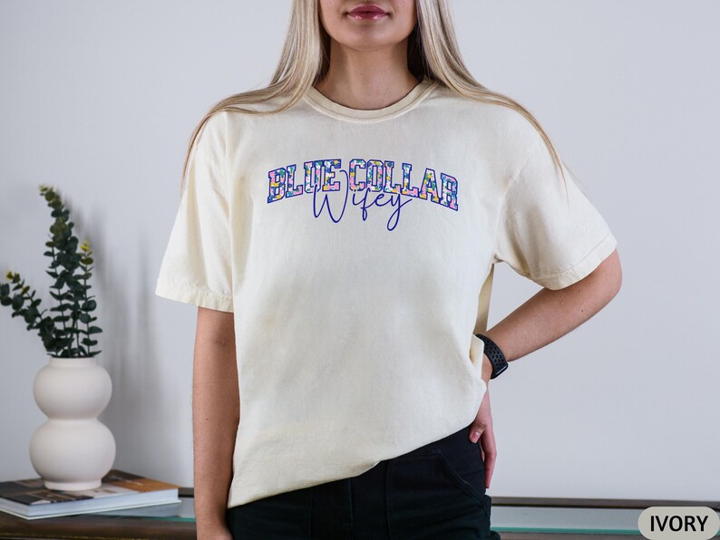 May include: Ivory t-shirt featuring the text "BLUE COLLAR WIFEY" in a floral design. The shirt has a relaxed fit and short sleeves. The text is in a blue and floral design.