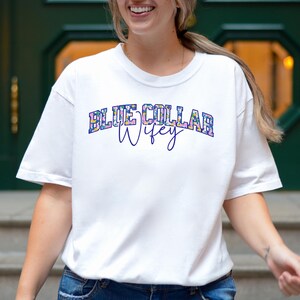May include: White t-shirt featuring the text "BLUE COLLAR Wifey" in a floral design. The shirt has a crew neck and is made of a soft fabric.