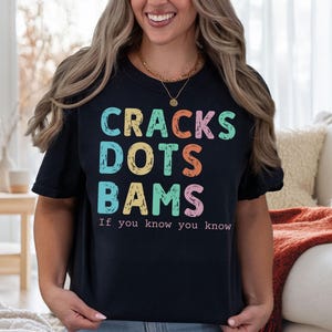 May include: Black t-shirt with the words "CRACKS DOTS BAMS" in colorful, block letters. Below the words is the phrase "If you know you know". The shirt is a casual style.