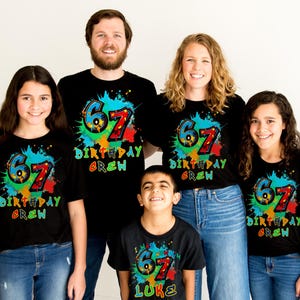 May include: A group of five people wearing matching black t-shirts. Each shirt features a colorful design with the number "67" and the words "Birthday Crew". The design includes splashes of blue, green, red, and yellow.