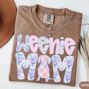 K&ouml;nnte beinhalten: Espressofarbenes T-Shirt mit dem Schriftzug "Weenie Mom" in einem floralen und Vichy-Muster. Eine Dackel-Illustration befindet sich in der Mitte. Eine silberne Halskette mit einem kleinen Anh&auml;nger befindet sich auf dem Shirt. Ein brauner Hut befindet sich in der oberen linken Ecke.
