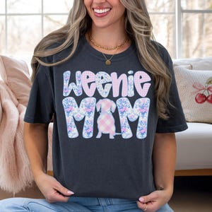 K&ouml;nnte beinhalten: Dunkelgraues T-Shirt mit den Worten "Weenie Mom" in einem floralen und Vichy-Design. Das Shirt zeigt eine Dackel-Illustration in der Mitte, ebenfalls mit einem Vichy-Muster. Das Shirt ist ein l&auml;ssiger, bequemer Stil.