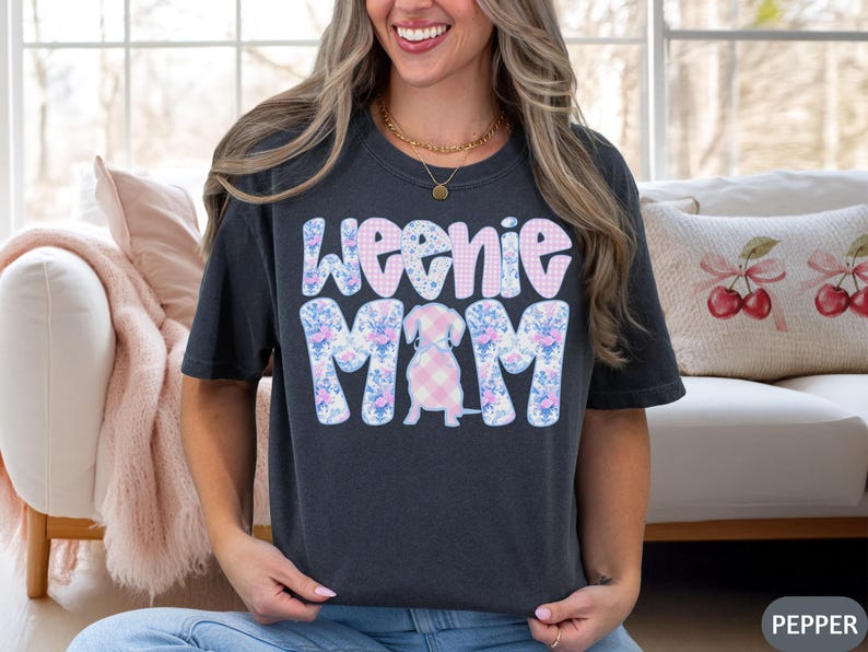 K&ouml;nnte beinhalten: Dunkelgraues T-Shirt mit den Worten "Weenie Mom" in einem floralen und Vichy-Design. Das Shirt zeigt eine Dackel-Illustration in der Mitte, ebenfalls mit einem Vichy-Muster. Das Shirt ist ein l&auml;ssiger, bequemer Stil.