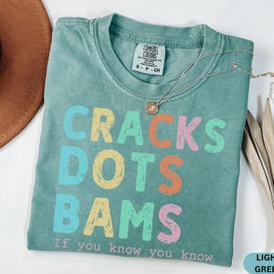 May include: Light green t-shirt with the words "CRACKS DOTS BAMS" in colorful letters. The text "If you know you know" is below the main text. A gold necklace and a brown hat are also in the image.