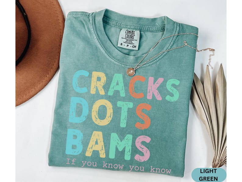 May include: Light green t-shirt with the words "CRACKS DOTS BAMS" in colorful letters. The text "If you know you know" is below the main text. A gold necklace and a brown hat are also in the image.