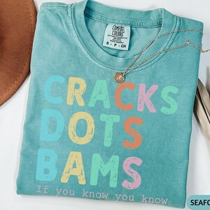 May include: Seafoam green t-shirt with the words "CRACKS DOTS BAMS" in colorful, distressed lettering. The phrase "If you know you know" is printed below. A gold necklace with a pendant rests on the shirt.