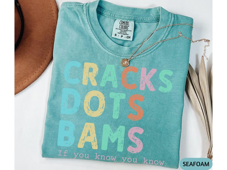 May include: Seafoam green t-shirt with the words "CRACKS DOTS BAMS" in colorful, distressed lettering. The phrase "If you know you know" is printed below. A gold necklace with a pendant rests on the shirt.