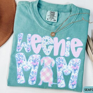 K&ouml;nnte beinhalten: T&uuml;rkisgr&uuml;nes T-Shirt mit dem Schriftzug "Weenie Mom" in einem floralen und Vichy-Muster. Eine Dackel-Grafik befindet sich in der Mitte. Eine goldene Halskette mit Anh&auml;nger befindet sich auf dem Shirt.