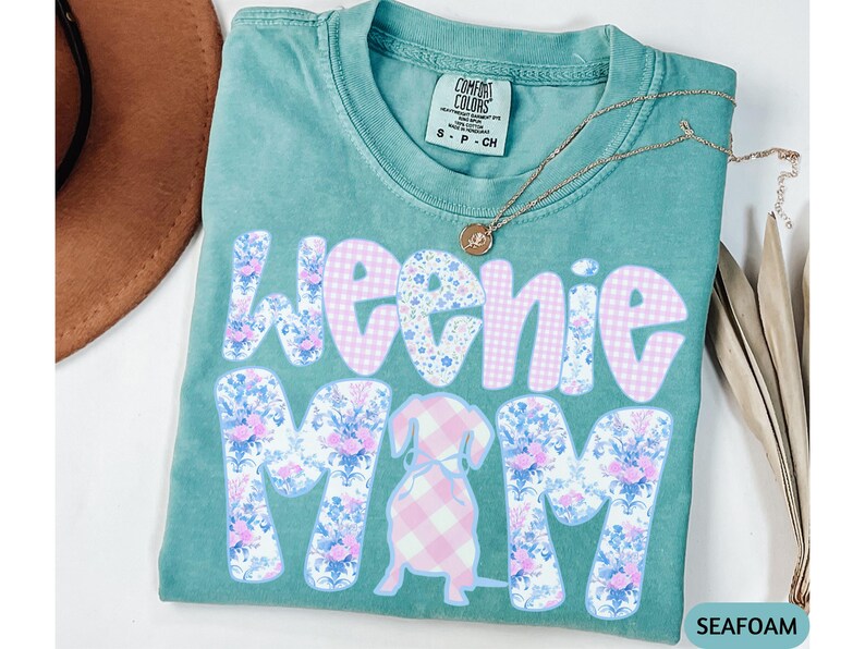 K&ouml;nnte beinhalten: T&uuml;rkisgr&uuml;nes T-Shirt mit dem Schriftzug "Weenie Mom" in einem floralen und Vichy-Muster. Eine Dackel-Grafik befindet sich in der Mitte. Eine goldene Halskette mit Anh&auml;nger befindet sich auf dem Shirt.