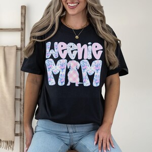 K&ouml;nnte beinhalten: Schwarzes T-Shirt mit dem Text "Weenie Mom" in einem floralen und karierten Design. Das Shirt zeigt eine Dackel-Hunde-Illustration. Das Wort "BLACK" befindet sich in einem schwarzen Rechteck unten rechts.