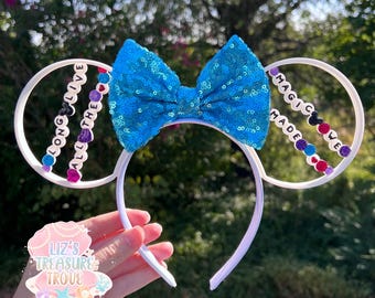 Peut inclure: Des oreilles de souris blanches avec un nœud en sequins bleus et des breloques perlées qui épellent "Long Live All The Magic We Made".