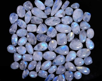 May include: A collection of polished moonstone gemstones in various shapes, including oval, teardrop, and rectangular. The stones display a milky white base with iridescent blue and silver flashes. The gemstones are arranged on a black background.