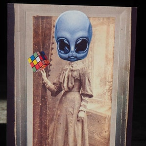 May include: A vintage-style print featuring a blue alien head superimposed on a sepia-toned portrait of a woman in a long dress. The alien holds a colorful Rubik's Cube. The print has a dark border and a distressed aesthetic.