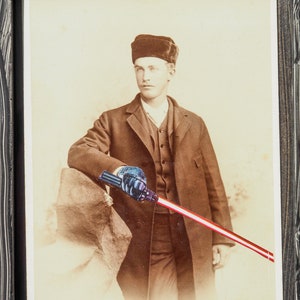 Peut inclure: Photographie sépia encadrée d'un homme en manteau et chapeau sombres, tenant un sabre laser rouge et blanc. La photo porte le texte "BANGS" et "SLEEPY EYE, MINN." en bas.