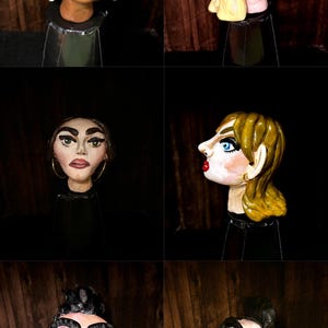 May include: A collection of six hand-painted ceramic sculptures of human heads. Each head features unique facial expressions, hairstyles, and accessories, including earrings and nose rings. The sculptures are set against a dark background.