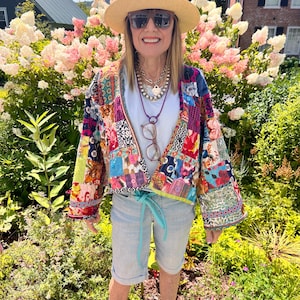 May include: A woman wearing a straw hat, sunglasses, and a colorful patchwork jacket. She is also wearing a white t-shirt, light blue shorts, and brown shoes. The background features lush greenery and pink and white flowers.