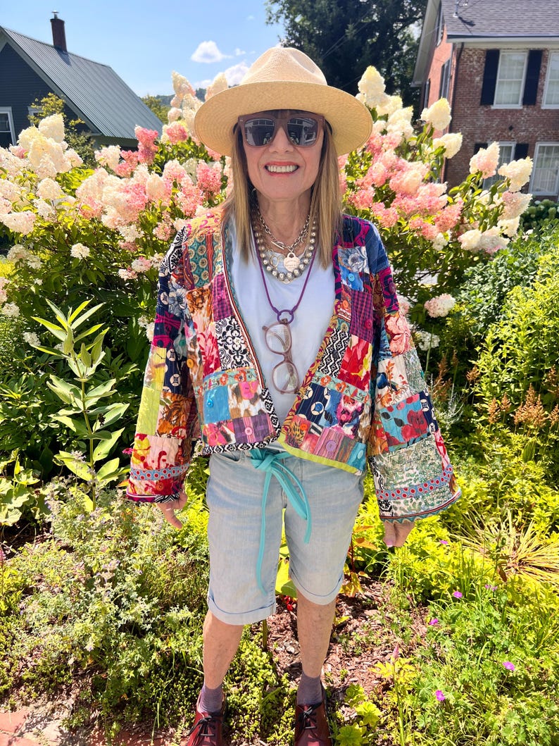 May include: A woman wearing a straw hat, sunglasses, and a colorful patchwork jacket. She is also wearing a white t-shirt, light blue shorts, and brown shoes. The background features lush greenery and pink and white flowers.