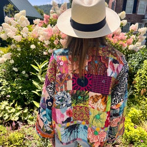 May include: A straw fedora hat with a black band. A colorful patchwork jacket with various patterns and colors, including floral and geometric designs. The jacket has button details and is worn outdoors.