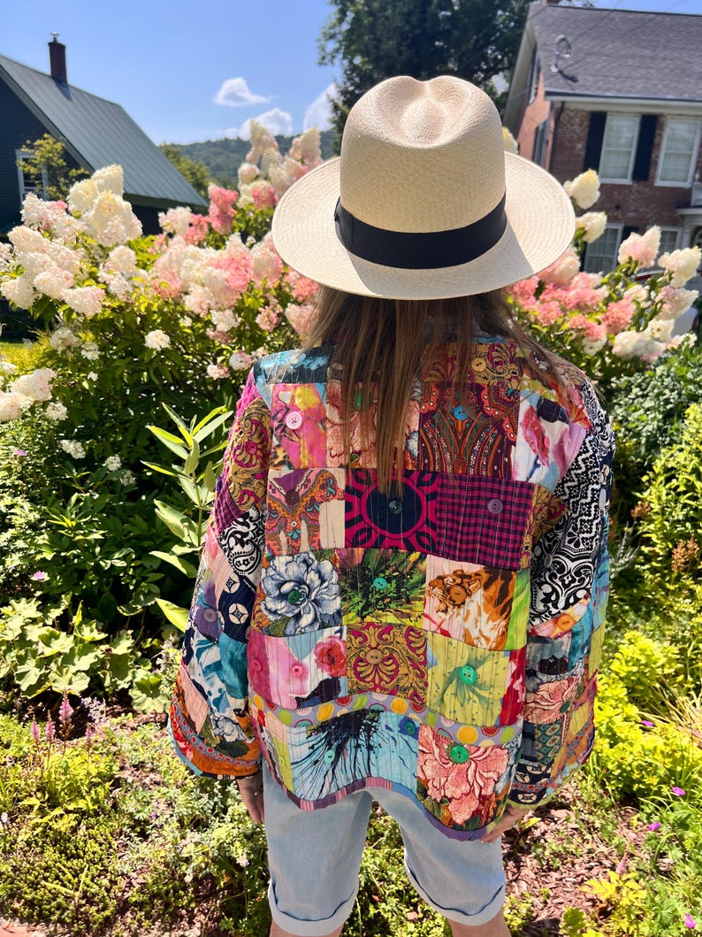 May include: A straw fedora hat with a black band. A colorful patchwork jacket with various patterns and colors, including floral and geometric designs. The jacket has button details and is worn outdoors.