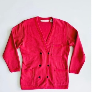 May include: A red cardigan with a v-neck, two pockets, and a button closure. The cardigan has a ribbed texture and black buttons.
