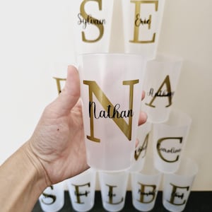 Peut inclure: Plusieurs gobelets en plastique transparents avec des lettres dorées. Un gobelet est tenu dans une main. Les gobelets portent des noms comme "Nathan", "Sylvain" et "Eric". Pour une fête ou un événement.