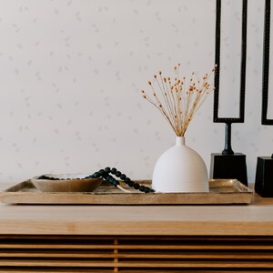 May include: A white ceramic vase with dried flowers sits on a wooden tray with a black beaded necklace and a gold bowl. Two black metal sculptures stand behind the tray.