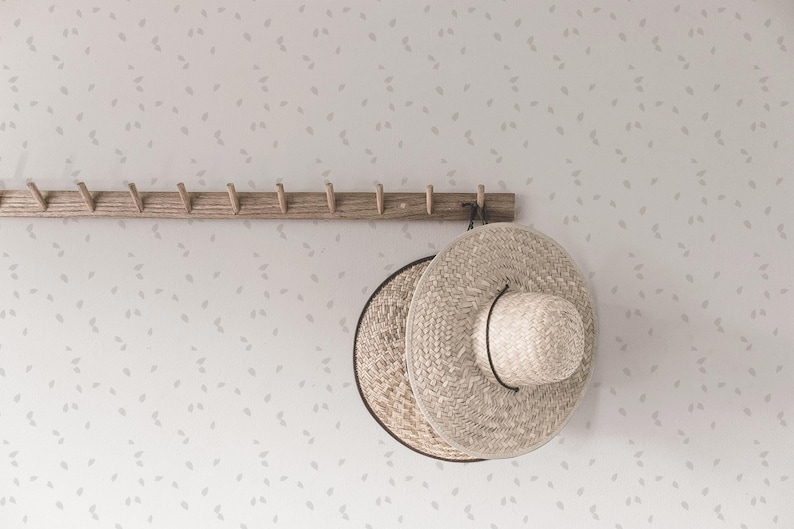 May include: Two woven straw hats hanging on a wooden rack with hooks against a white wall with a subtle pattern of gray leaves.