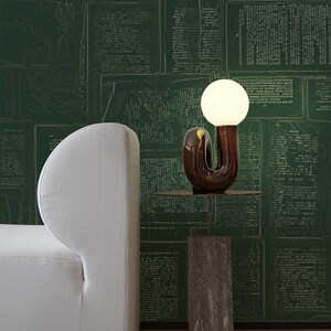 May include: A white couch with a curved back sits in front of a green wall with a white patterned design. A brown ceramic lamp with a white globe sits on a brown side table.