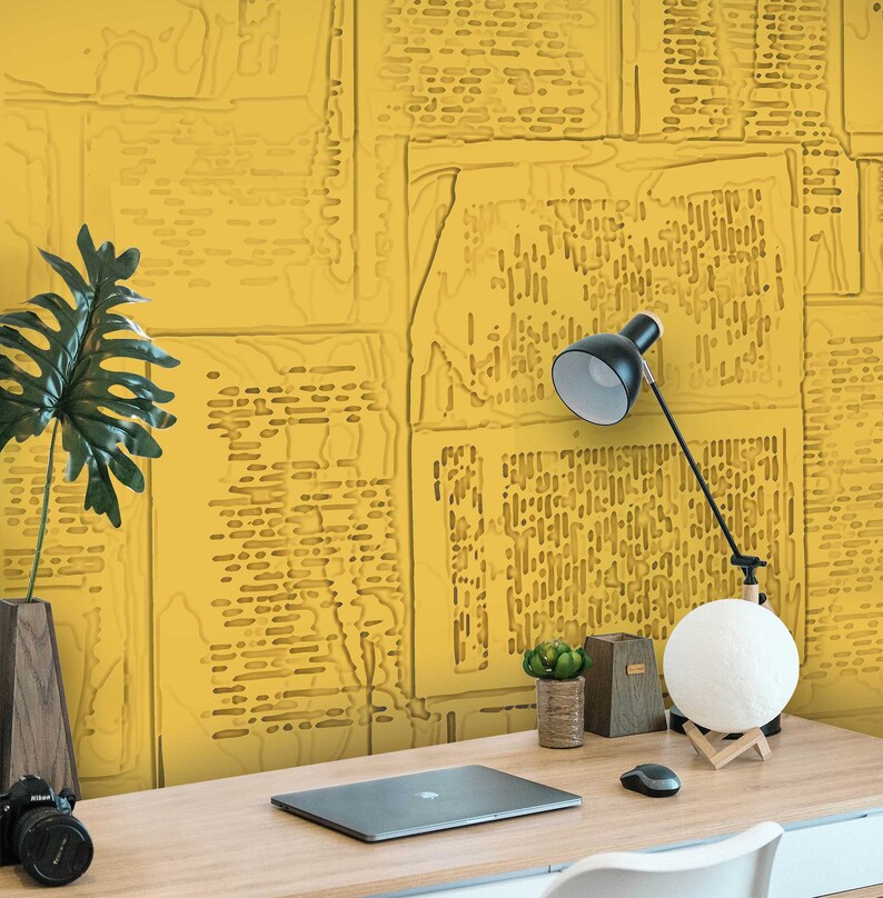 May include: A yellow desk with a laptop, a lamp, a moon lamp, and two small potted plants. The wall behind the desk is covered in a yellow wallpaper with a pattern of overlapping newspaper clippings.