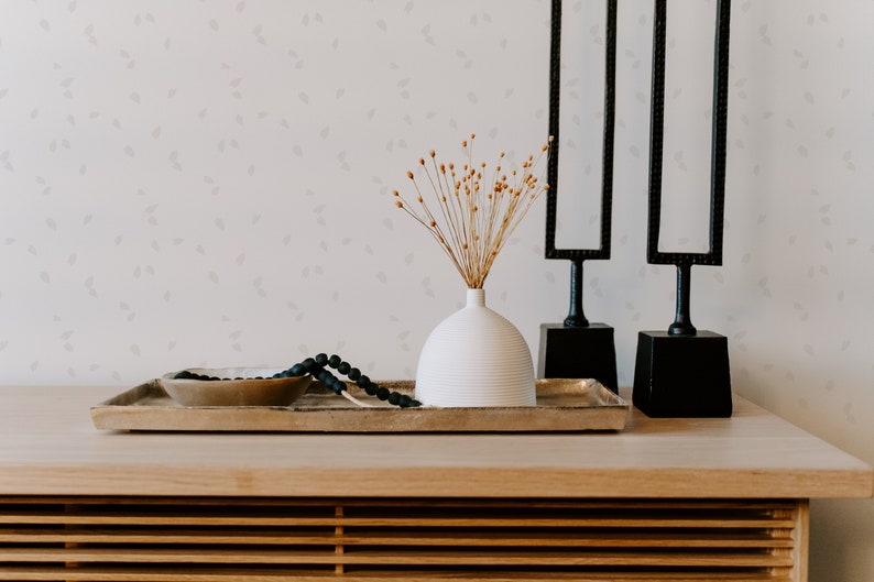 May include: A white ceramic vase with dried flowers sits on a wooden tray with a black beaded necklace and a gold bowl. Two black metal sculptures stand behind the tray.