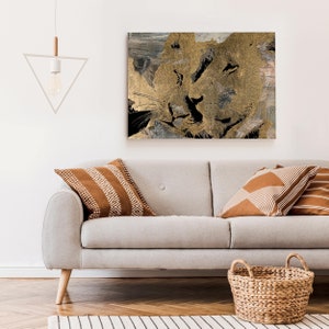 Puede incluir: Una pintura abstracta de dos leones enfrentados, realizada en oro y negro. La pintura está colgada en una pared blanca sobre un sofá gris claro con cojines marrones.
