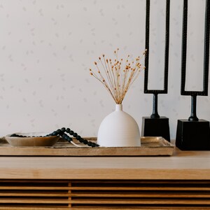 May include: A white ceramic vase with dried flowers sits on a wooden tray with a black beaded necklace and a gold bowl. Two black metal sculptures with rectangular frames stand behind the tray.
