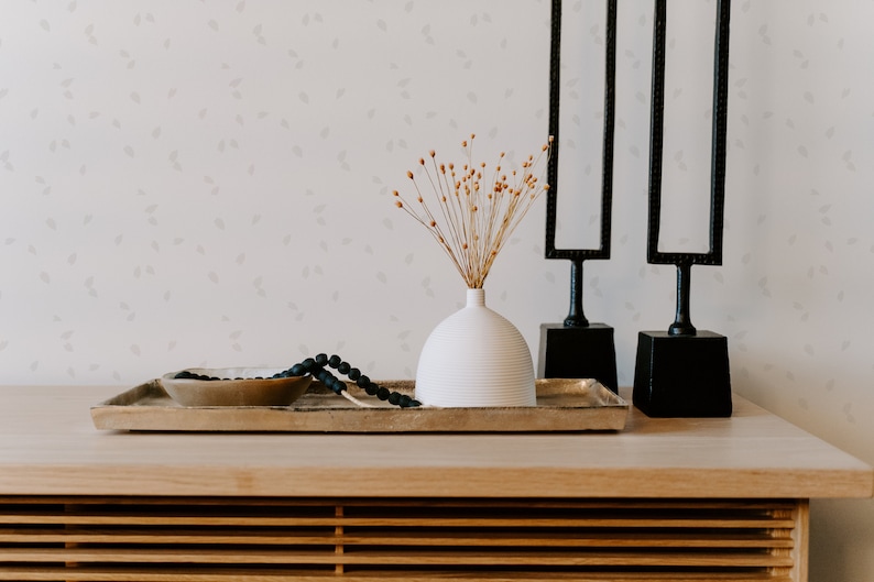 May include: A white ceramic vase with dried flowers sits on a wooden tray with a black beaded necklace and a gold bowl. Two black metal sculptures with rectangular frames stand behind the tray.