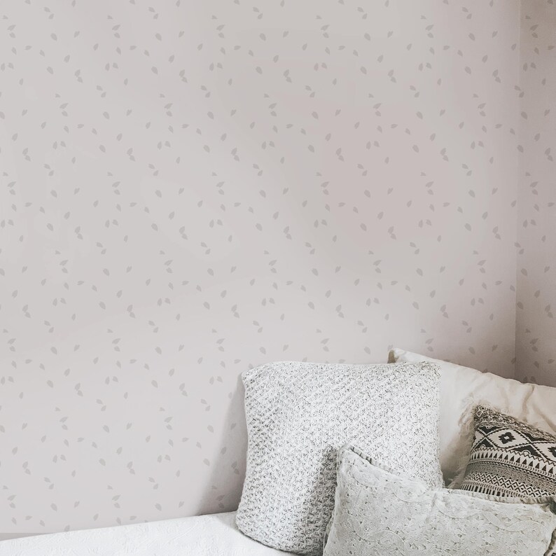 May include: A light pink wall with a subtle pattern of small gray leaves. A white bed with a gray knitted throw pillow and two white pillows is in the corner of the room.