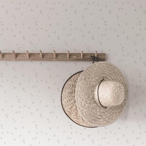 May include: Two woven straw hats hanging on a wooden rack with hooks against a white wall with a subtle pattern of gray leaves.