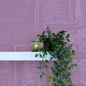 May include: A green glass candle holder with a lit candle sits on a white shelf with a potted green plant hanging over the edge. The background is a pink wall with a white patterned design.
