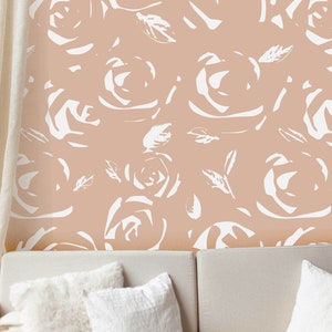 May include: A peach-colored wall with a repeating pattern of white rose illustrations. The roses and leaves are stylized with brushstroke-like details. Below, a white sofa with fluffy and textured pillows.